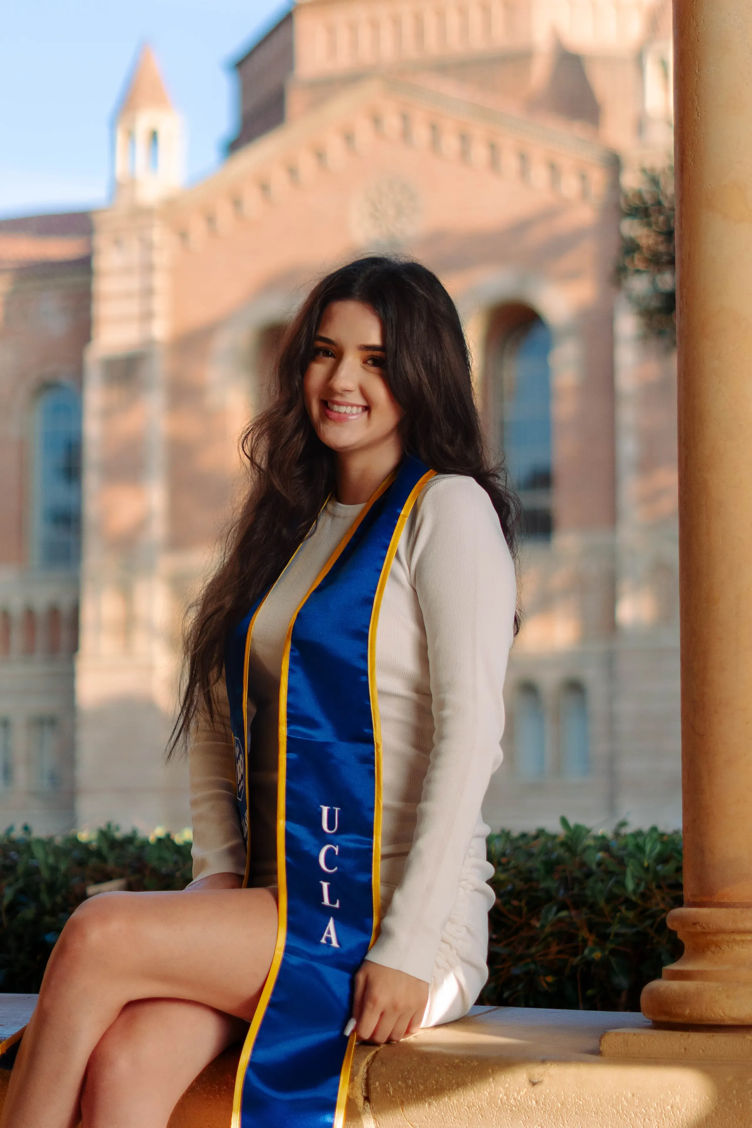 UCLA graduate seated beneath a campus arcade in a blue and gold commencement stole