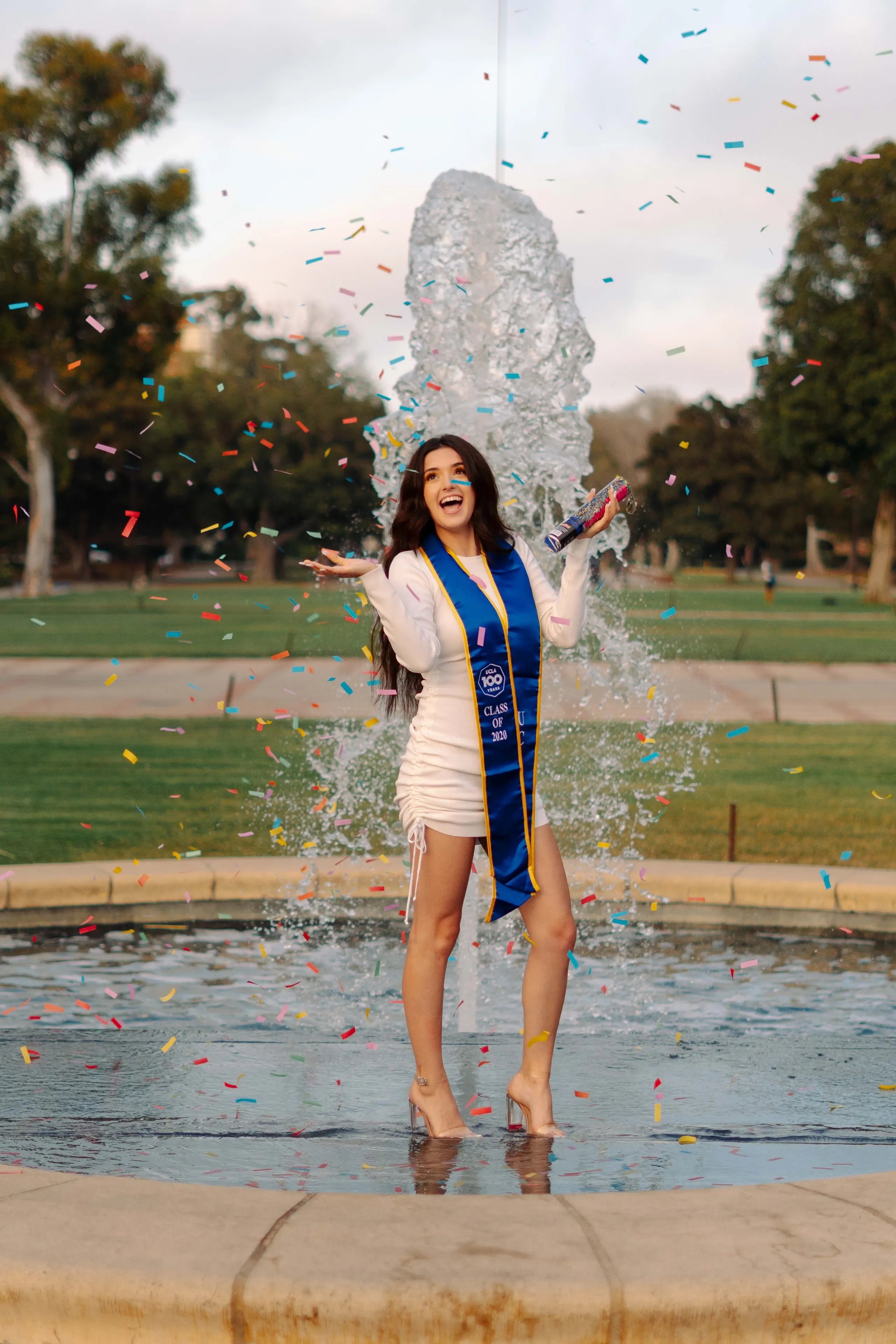 UCLA graduate celebrating in a fountain as colorful confetti falls